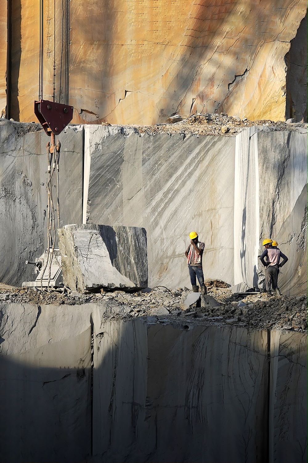 Workers at the foot of a towering marble quarry wall, a freshly cut block and crane beside them